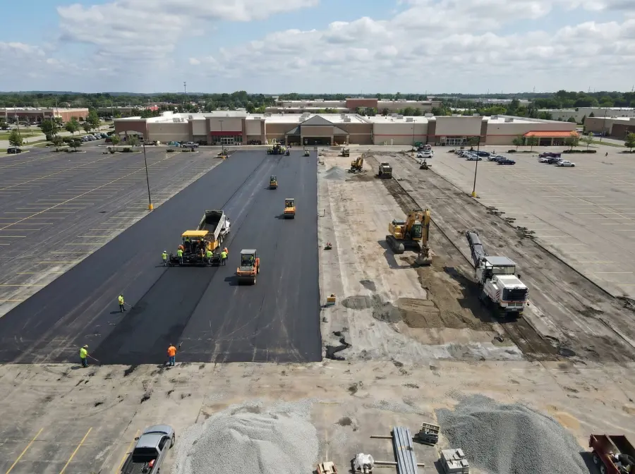 Aerial view of a large commercial parking lot project, showing side-by-side asphalt paving and heavy-duty reconstruction operations on a retail site.